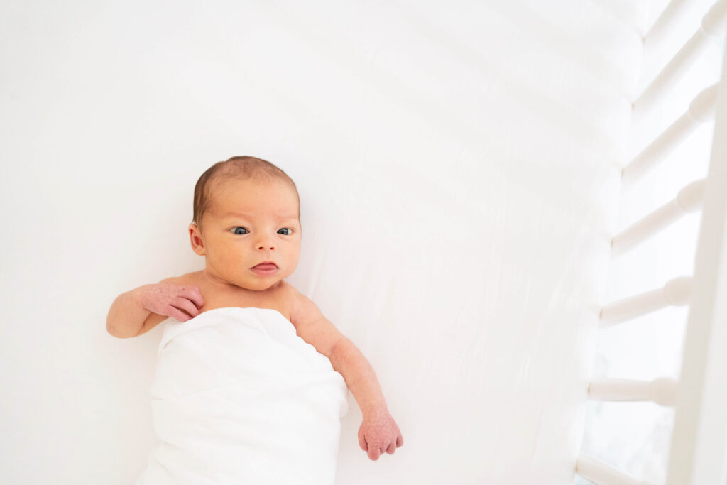 Newborn baby in crib looking up with white swaddle on white sheet. 