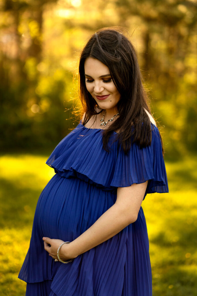 Pregnant woman in blue dress looking at stomach and smiling against sunset.