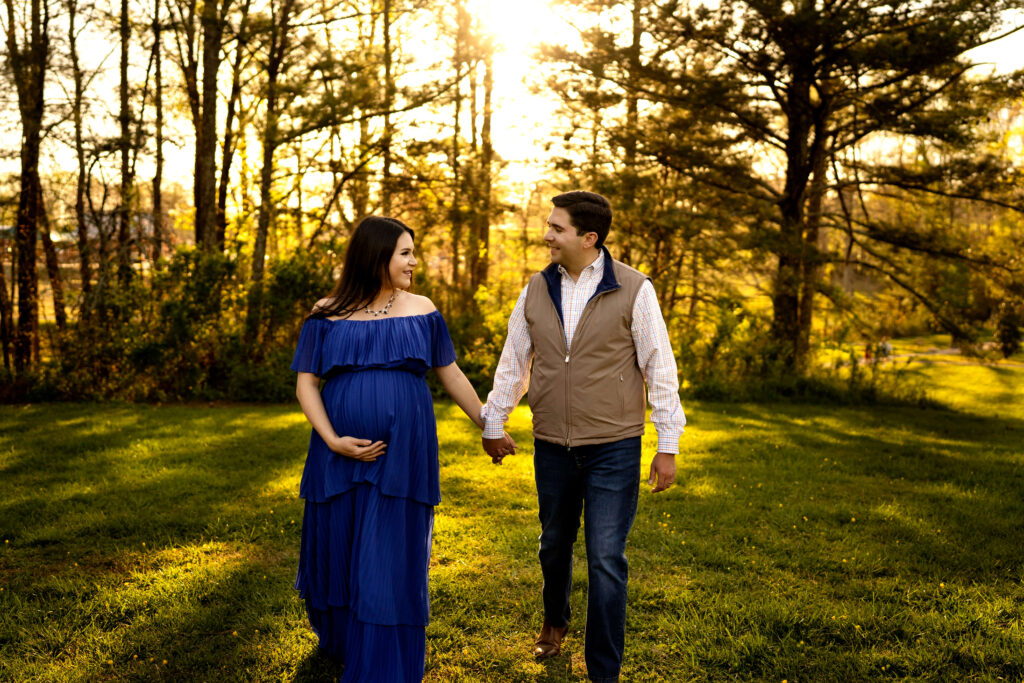 Pregnant woman in blue dress looking at husband while walking across field during sunset.