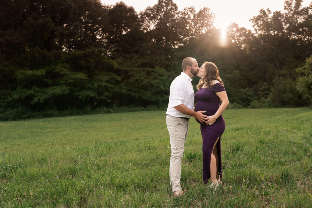 Standing in a sunset field, husband kissing pregnant wife and purple maternity dress. 