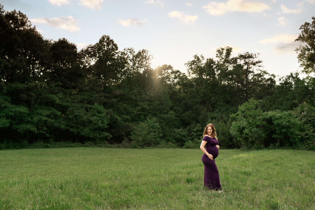 Pregnant woman in field posing in purple dress. 