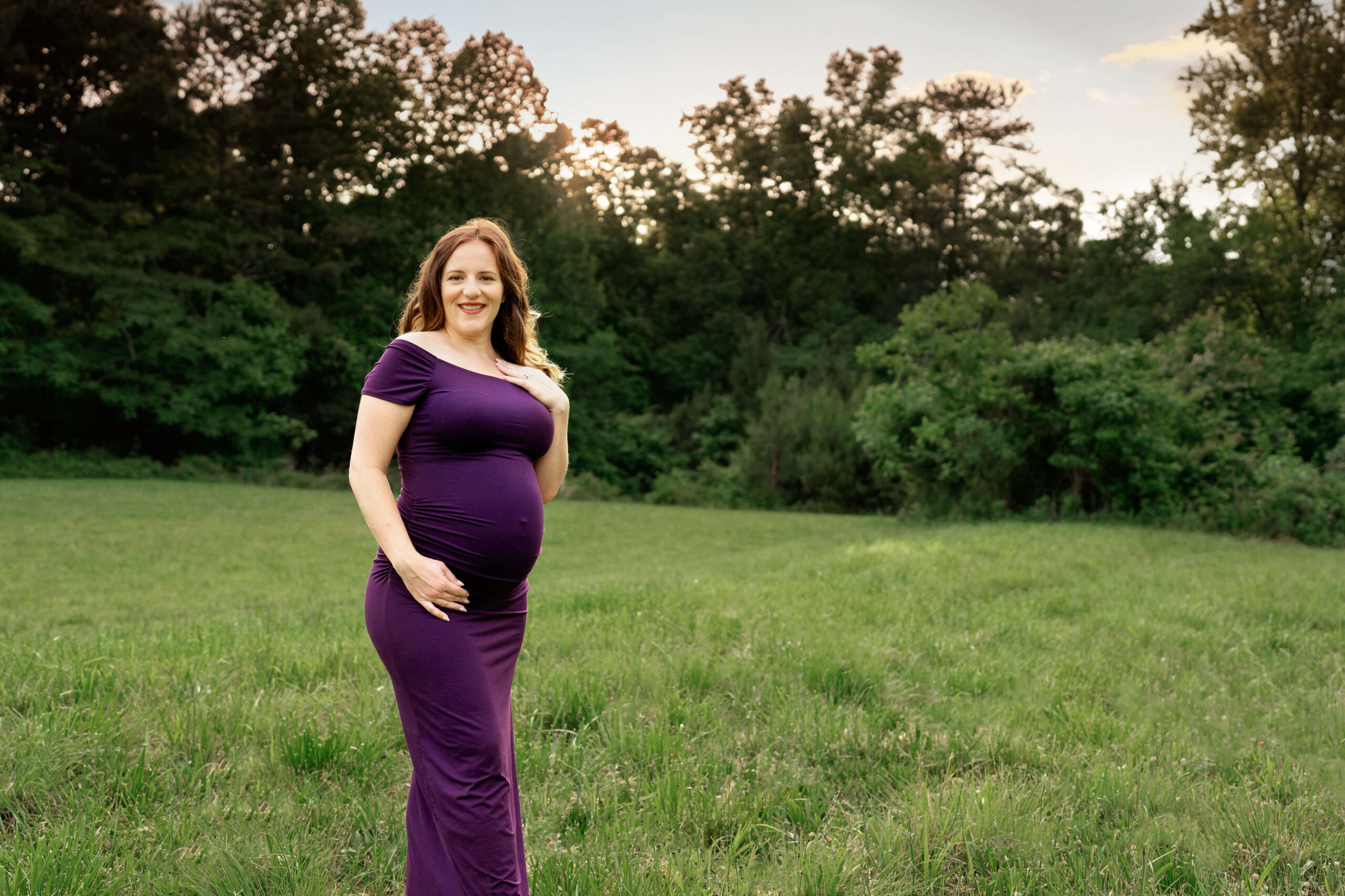 Pregnant woman in field in purple dress smiling at camera.