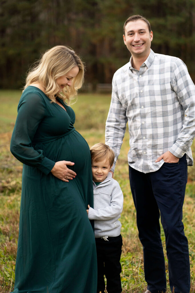 Pregnant woman in green dress looking down at young son who is hugging her while husband smiles at camera.