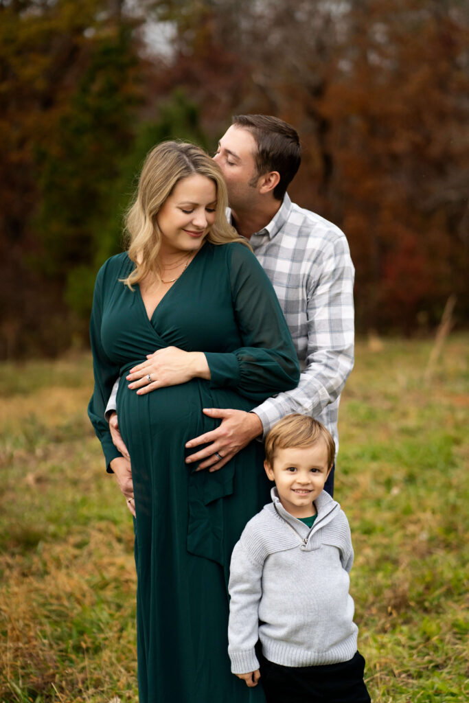 Pregnant woman in green dress with her eyes closed while husband kisses her from behind and young son looks at camera. 