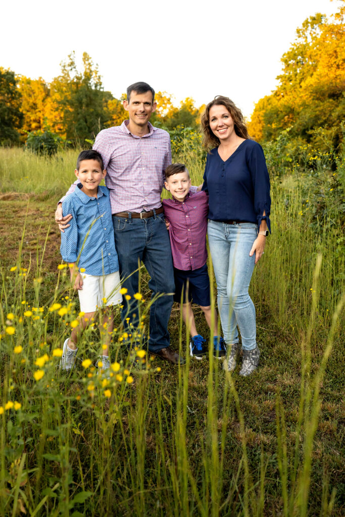 Mother, father and two young sons standing in a field of tall grass, looking at the camera and smiling. 