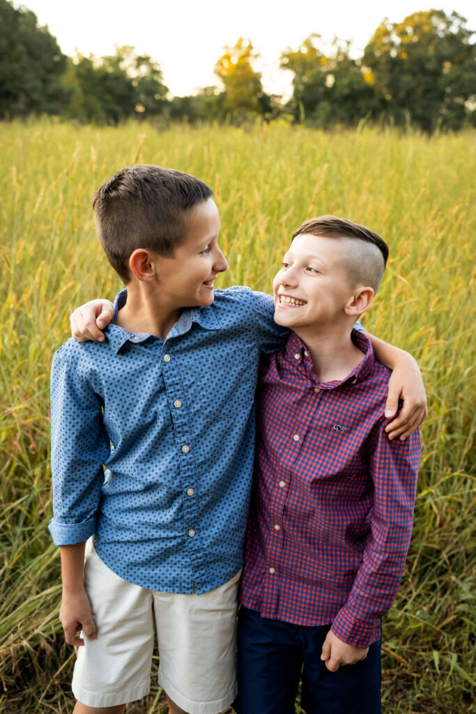 Two brothers standing with their arms around each other and smiling at each other in a field of tall grass. 