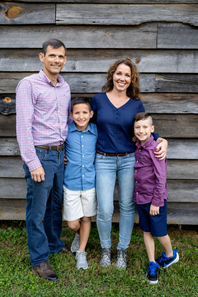 Mother, father and two sons standing against wooden building, looking at camera and smiling. 