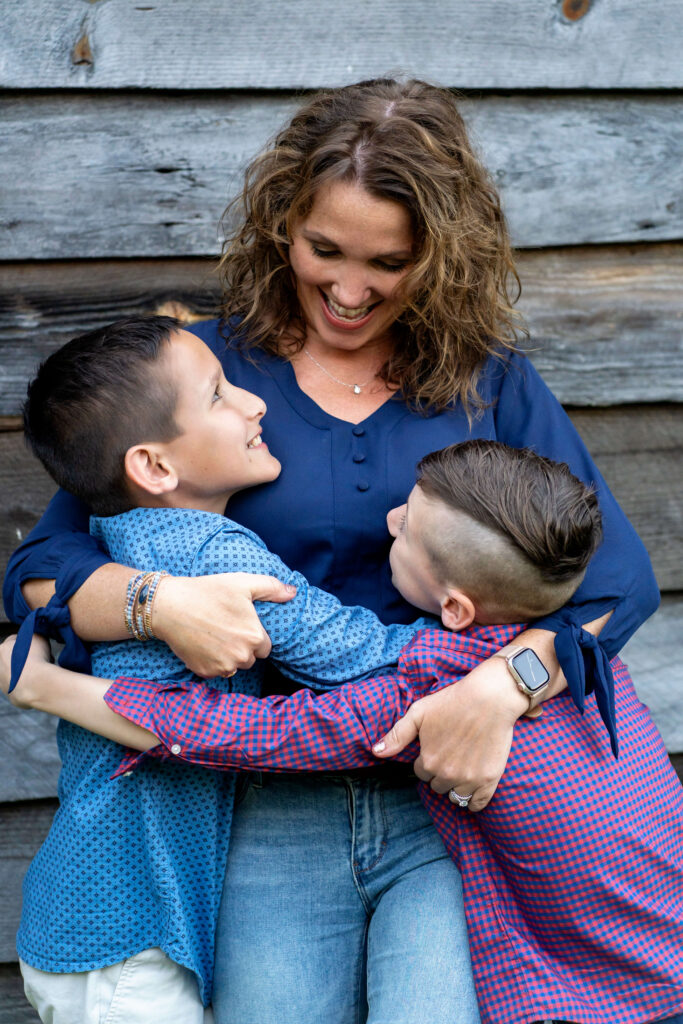 Mother hugging two young sons while standing in front of wooden building. 