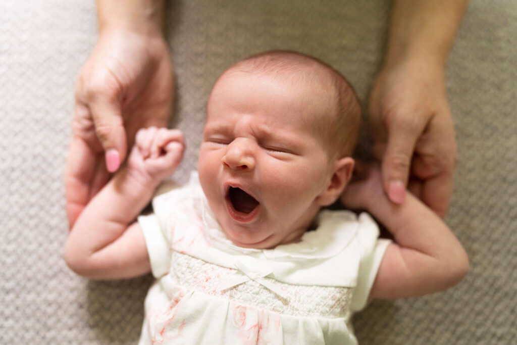 Newborn girl yawning while laying on a chair and mother holds her hands